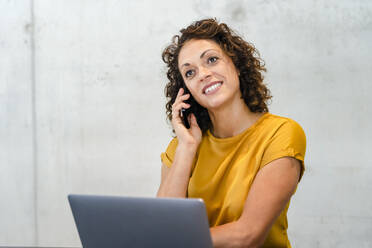 Smiling businesswoman with laptop hand on chin in front of wall - DIGF16799