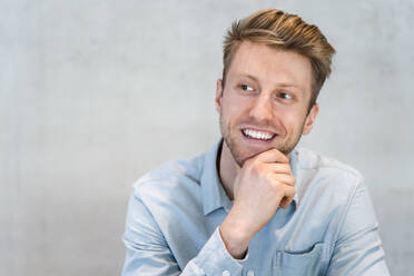 Smiling young businessman with hand on chin in front of wall - DIGF16835