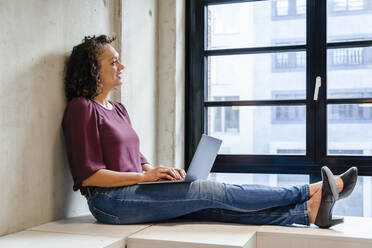 Smiling businesswoman with laptop looking through window in office - DIGF16910