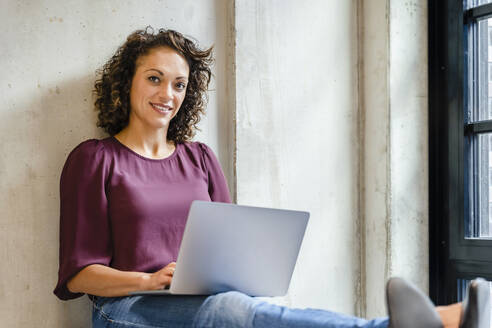 Lächelnde Geschäftsfrau mit Laptop auf dem Schoß im Büro - DIGF16913