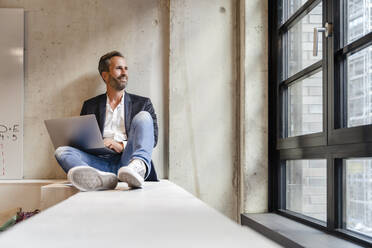 Thoughtful businessman with laptop looking out office window - DIGF16915