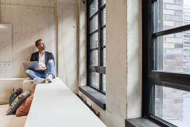 Businessman with laptop sitting by window - DIGF16916