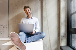 Lächelnder junger Geschäftsmann mit Laptop, der am Bürofenster sitzt - DIGF16920