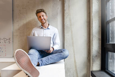 Smiling young businessman with laptop sitting at office window - DIGF16920