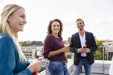 Lächelnde Geschäftsfrau mit Alkoholflasche und Kollegen auf dem Dach - DIGF16927