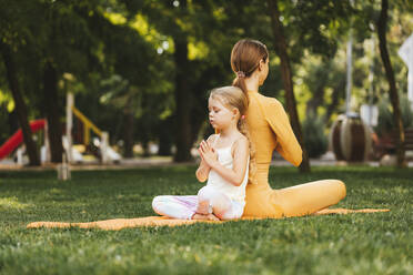 Girl with hands clasped meditating while sitting back to back of mother in park - OYF00631