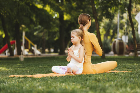 Mädchen mit verschränkten Händen meditiert mit dem Rücken zu ihrer Mutter im Park sitzend - OYF00631
