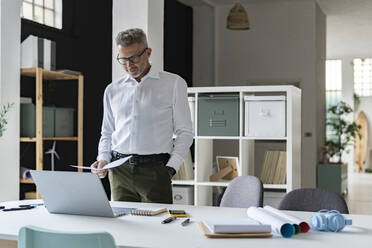 Geschäftsmann hält ein Dokument in der Hand und schaut auf einen Laptop im Büro - GIOF13928