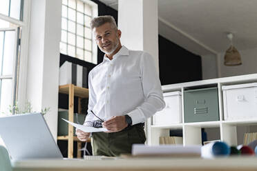 Smiling businessman with document and eyeglasses at desk - GIOF13929