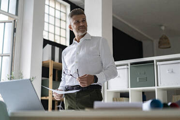 Businessman holding document and eyeglasses in office - GIOF13930