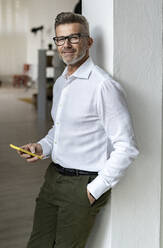 Lächelnder Geschäftsmann mit Smartphone an der Wand lehnend im Büro - GIOF13933