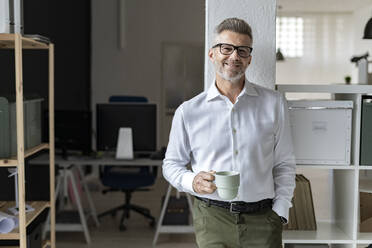 Smiling businessman with coffee mug leaning on architectural column - GIOF13941