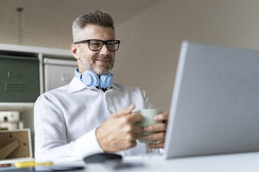 Smiling businessman with coffee mug on video conference through laptop in office - GIOF13945