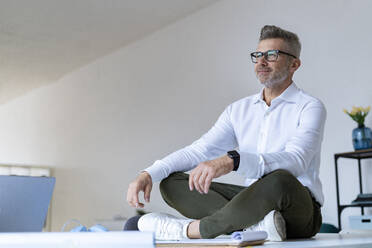 Geschäftsmann sitzt im Schneidersitz nachdenklich auf dem Schreibtisch im Büro - GIOF13952