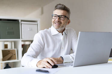 Smiling businessman with laptop contemplating in office - GIOF13956