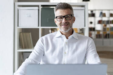 Businessman with hair stubble smiling in front of laptop at office - GIOF13957