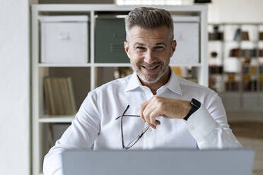 Smiling businessman holding eyeglasses at office - GIOF13959