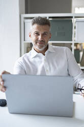 Lächelnder Geschäftsmann sitzt mit Laptop am Schreibtisch - GIOF13960