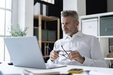 Thoughtful businessman holding eyeglasses looking at laptop in office - GIOF13961