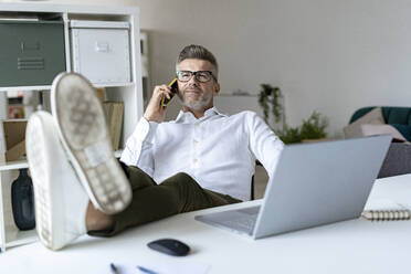 Geschäftsmann mit hochgelegten Füßen, der am Schreibtisch mit seinem Smartphone telefoniert - GIOF13966