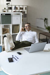 Lächelnder Geschäftsmann mit hochgelegten Füßen, der im Büro mit einem Mobiltelefon spricht - GIOF13967