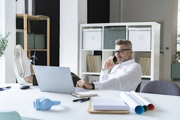 Businessman talking on smart phone while using laptop in office - GIOF13968