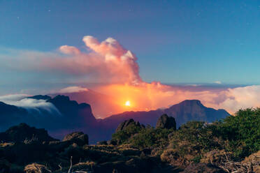 Nächtliche Landschaft mit einem ausbrechenden Vulkan im Hintergrund und einem Wolkenmeer, das die Berge eines bewachsenen und felsigen Berges bedeckt. Vulkanausbruch auf der Cumbre Vieja in La Palma, Kanarische Inseln, Spanien, 2021 - ADSF31275
