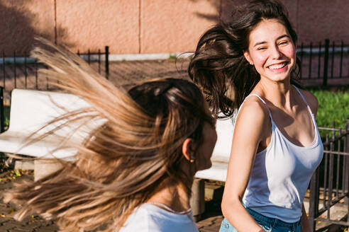 Fröhliche beste Freundinnen mit fliegenden braunen Haaren lachen, während sie an einem sonnigen Tag Zeit in der Stadt verbringen - ADSF31278