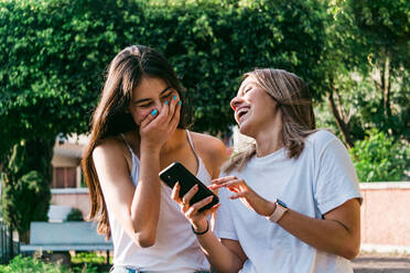 Smiling teen covering mouth while interacting with excited best female friend with cellphone in sunny town - ADSF31279