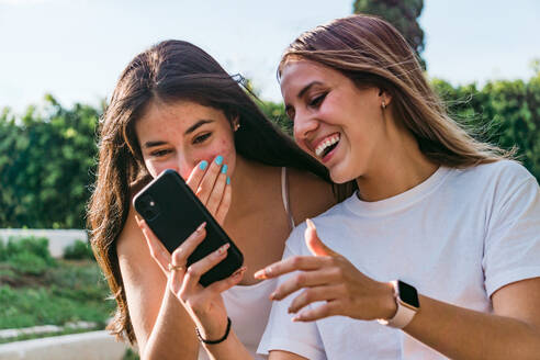 Lächelnder Teenager, der den Mund bedeckt, während er mit seiner aufgeregten besten Freundin mit dem Handy in einer sonnigen Stadt interagiert - ADSF31280