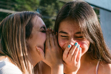 Fröhlicher Teenager, der sich den Mund zuhält, während er seiner besten Freundin mit geschlossenen Augen im Sonnenlicht etwas ins Ohr flüstert - ADSF31281