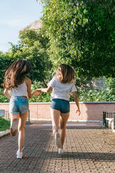 Back view of anonymous best female friends holding hands while running on tiled walkway in town - ADSF31282