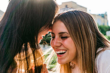 Cheerful teen covering mouth while whispering in ear of best female friend with closed eyes in sunlight - ADSF31283