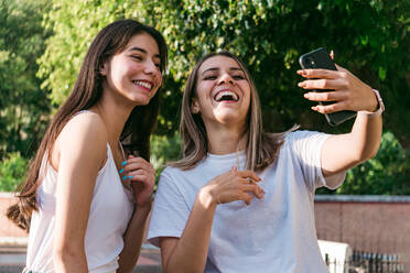 Cheerful female teenager near best friend with acne taking self portrait on cellphone in sunlight - ADSF31284