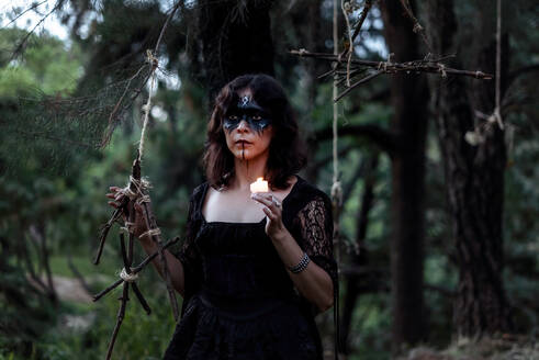 Mysteriöse Hexe in schwarzem Kleid und mit leuchtender Kerze, die im düsteren Wald steht und wegschaut - ADSF31320