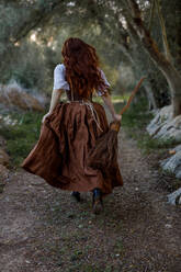 Back view of unrecognizable witch in dress and with broomstick running along path in autumn forest - ADSF31322