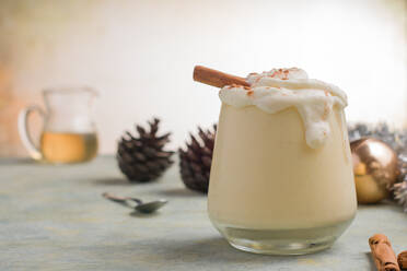 Glas Milchpunsch mit Zimtpulver auf geschlagenem Eiweiß gegen Tannenzapfen am Weihnachtstag auf hellem Hintergrund - ADSF31323