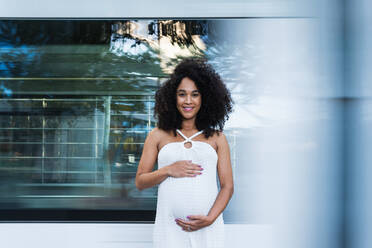 Charmante schwangere Afroamerikanerin mit Afro-Locken im weißen Kleid, die in der Nähe einer reflektierenden Wand steht und fröhlich in die Kamera lächelt - ADSF31332