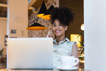 Positive afroamerikanische Frau mit Afrofrisur, die am Tisch mit Netbook und einer Tasse Kaffee mit einem Handy spricht - ADSF31337