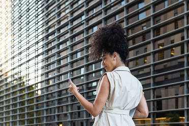 Rückenansicht einer jungen Afroamerikanerin mit lockigem Haar, die auf ihr Handy schaut, während sie ein Netbook an einem modernen Gebäude vorbei trägt - ADSF31342