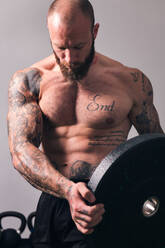 Sportler mit muskulösem Körper und nacktem Oberkörper, der mit einer schweren Gewichtsplatte in einem Fitnessstudio steht - ADSF31350