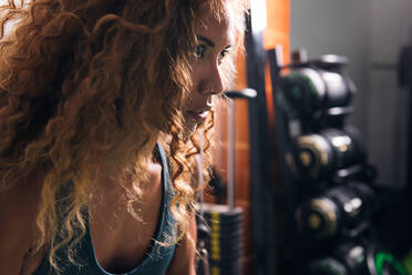 Seitenansicht der Ernte konzentrierte Sportlerin mit langen lockigen Haaren Blick nach vorne gegen Hanteln im Fitnessstudio in der Tageszeit - ADSF31351