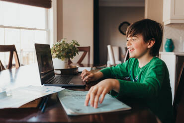 Happy boy doing on school work online on computer at kitchen table. - CAVF94946
