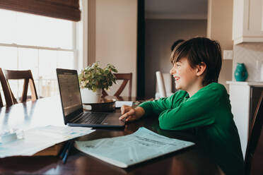 Happy boy doing on school work online on computer at kitchen table. - CAVF94948