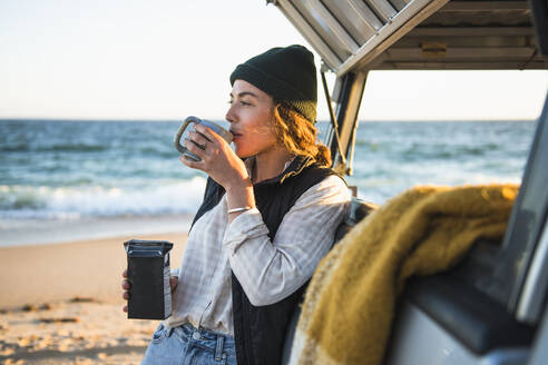 Junge Frau, die ein Getränk in einem Becher genießt, während sie allein im Auto am Strand campiert - CAVF94969