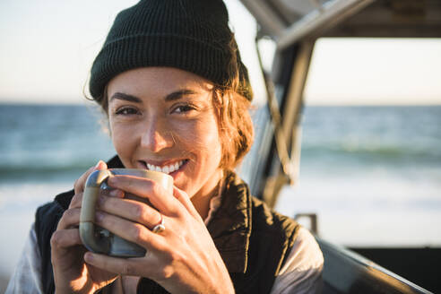 Young woman enjoying drink in mug while beach car camping alone - CAVF94970