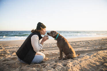 Junge Frau, die ein Getränk aus einem Becher genießt, während sie mit ihrem Hund am Strand zeltet - CAVF94972