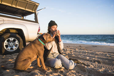 Junge Frau, die ein Getränk aus einem Becher genießt, während sie mit ihrem Hund am Strand zeltet - CAVF94973