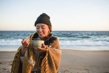 Junge Frau genießt eine Schüssel Suppe beim Camping mit dem Auto am Strand im Herbst - CAVF94974