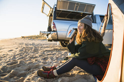 Junge Frau genießt Kaffee im Reisebecher beim Camping am Strand - CAVF94975